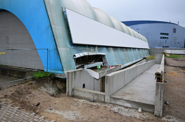 Das Bild zeigt eine Baustelle an der Längsseite der ehemaligen Eishalle: Vor der blau-weißen Hallenwand sind neue Betonrampen und Betonwände zu sehen. Hier entsteht ein barrierefreier, rollstuhlgerechter Zugang zum Gebäude. Im Hintergrund ist ein weiteres großes Hallengebäude erkennbar.