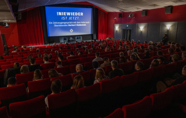 Das Foto zeigt eine Aufnahme aus einem Kinosaal aus der Perspektive der hinteren Reihen. Die Sitze haben rote Samtbezüge. Auch die Wände und die Vorhänge neben der Leinwand sind rot. Die Bühne vor der Leinwand ist mit zwei grauen Sesseln und einem Tisch mit Wasserflaschen bestückt. Im Saal sitzen mehrere Hundert Menschen. Auf der Leinwand steht auf blauem Hintergrund: „Nie wieder ist jetzt. Ein Zeitzeugengespräch mit dem Holocaust-Überlebenden Herbert Rubinstein.“  Darunter ist das Symbol der HSV-Raute.