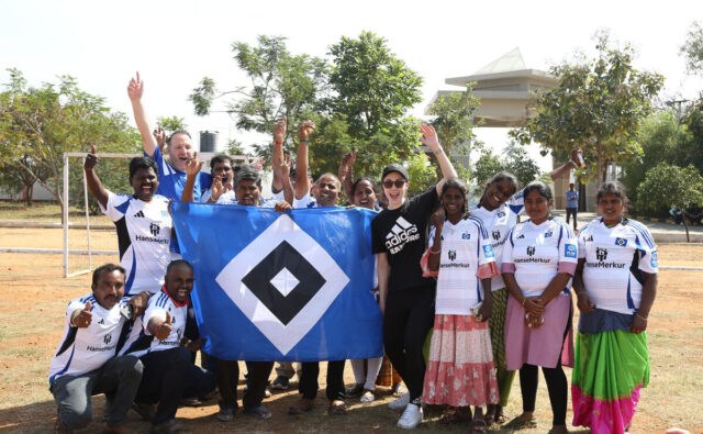 Das Bild zeigt eine Gruppe von 15 Menschen auf einem Fußballplatz. Sie jubeln. Die meisten von ihnen tragen das Heimtrikot des HSV aus der Saison 2024/25. Drei Personen halten eine HSV-Flagge in die Luft.