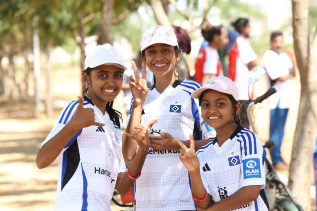 Das Foto zeigt drei Kinder, die in die Kamera lachen. Sie alle tragen helle Schirmmützen und das Heimtrikot des HSV aus der Saison 2024/25.