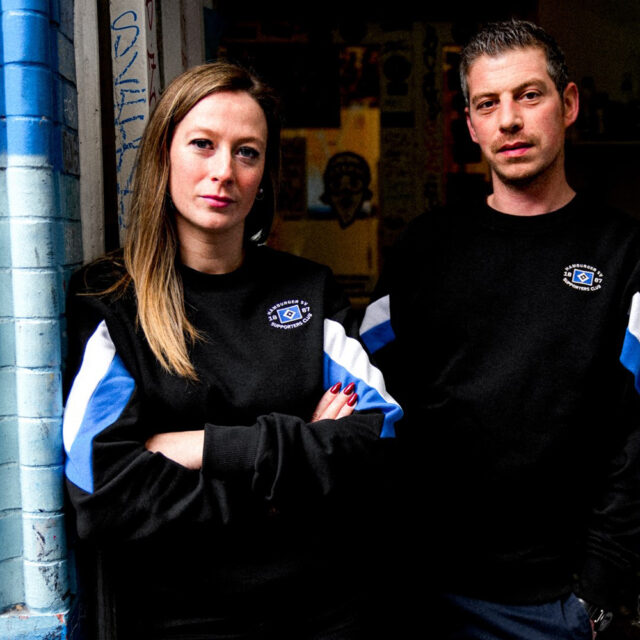 Auf dem Foto stehen eine Frau und ein Mann nebeneinander. Beide tragen einen Pullover aus der Merchandising-Kollektion exklusiv für Mitglieder des Supporters Clubs. Die Pullover sind schwarz mit großen weißen und blauen Streifen an den Armen. Die Frau lehnt an einer Wand. Beide schauen in die Kamera.