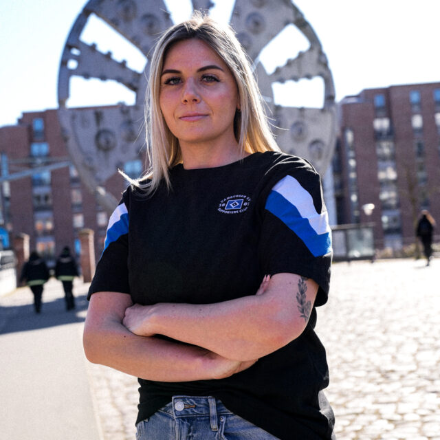 Auf dem Foto steht eine Frau mit verschränkten Armen auf einem öffentlichen Platz. Sie trägt ein T‑Shirt aus der Merchandising-Kollektion, welche exklusiv für Mitglieder des Supporters Clubs ist. Das T‑Shirt ist schwarz mit großen weißen und blauen Streifen an den Armen.