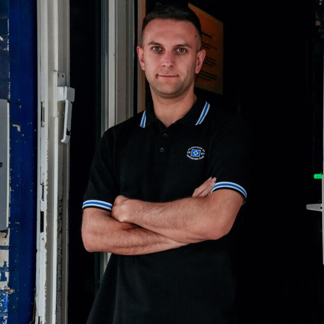 Auf dem Foto steht ein Mann mit verschränkten Armen neben einer Wand. Er trägt ein Poloshirt aus der Merchandising-Kollektion, welche exklusiv für Mitglieder des Supporters Clubs ist. Das Poloshirt ist schwarz und hat weiße sowie blaue Streife an den Ärmeln und dem Kragen.