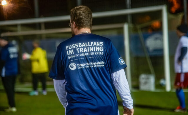 Das Foto zeigt einen Mann auf einem Fußballfeld, der von hinten zu sehen ist. Er trägt ein blaues Trikot mit der Aufschrift „Fußballfans im Training – gemeinsam gegen Krebs“ sowie dem Logo der Deutschen Krebshilfe und des HSV Supporters Clubs. Im Hintergrund sind weitere Personen beim Training und ein Tor zu erkennen. Die Szene spielt am Abend unter Flutlicht.