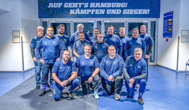Das Foto zeigt die Gruppe Fußballfans im Training (FFIT) des Hamburger SV. Die Männer tragen blaue Trainingsanzüge mit dem HSV-Logo und posieren in zwei Reihen in der hell beleuchteten MizedZone. Über ihnen hängt ein Schild mit der Aufschrift „Auf geht’s Hamburg! Kämpfen und siegen!“. Die Stimmung wirkt motiviert und gemeinschaftlich.