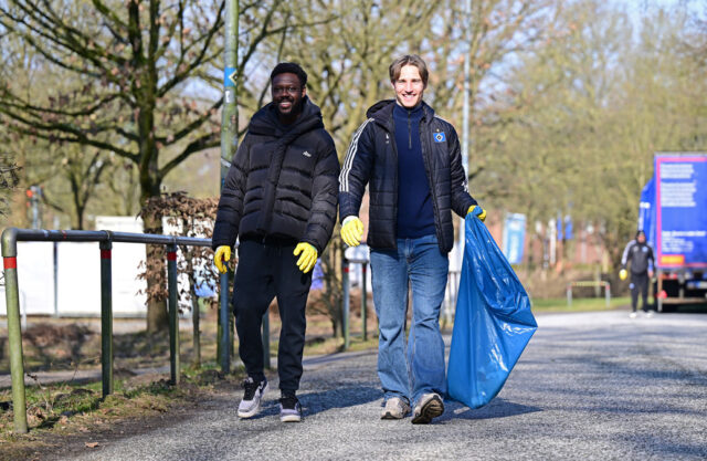 Das Bild zeigt zwei junge Männer, die auf die Kamera zugehen. Sie sind draußen. Sie tragen beide gelbe Plastikhandschuhe. Der rechte Mann trägt zusätzlich einen blauen Müllsack.
