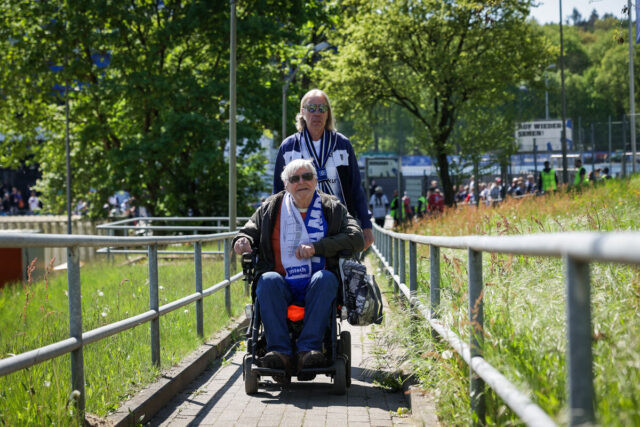 Das Bild zeigt die beiden Männer des Titelbildes auf dem Weg ins Volksparkstadion. Der Mann im Rollstuhl fährt vor dem Mann ohne Rollstuhl am Volksparkstadion eine Rampe hinauf. Es ist sonnig. Beide Männer tragen Sonnenbrillen.