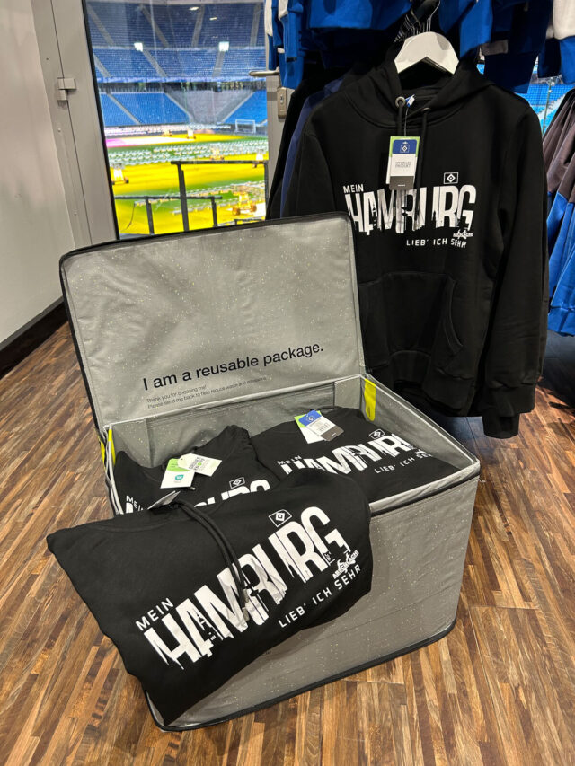 Auf dem Bild sind mehrere schwarze HSV-Hoodies mit dem weißen Aufdruck „Mein Hamburg lieb’ ich sehr“ zu sehen. Die Pullover liegen ordentlich gefaltet in der wiederverwendbaren Verpackungsbox mit der Aufschrift „I am a reusable package“ von der hey circle GmbH. Einer der Hoodies hängt daneben auf einem Kleiderbügel. Im Hintergrund ist durch das Fenster das Innere des Volksparkstadions mit dem Spielfeld und den Tribünen zu erkennen.