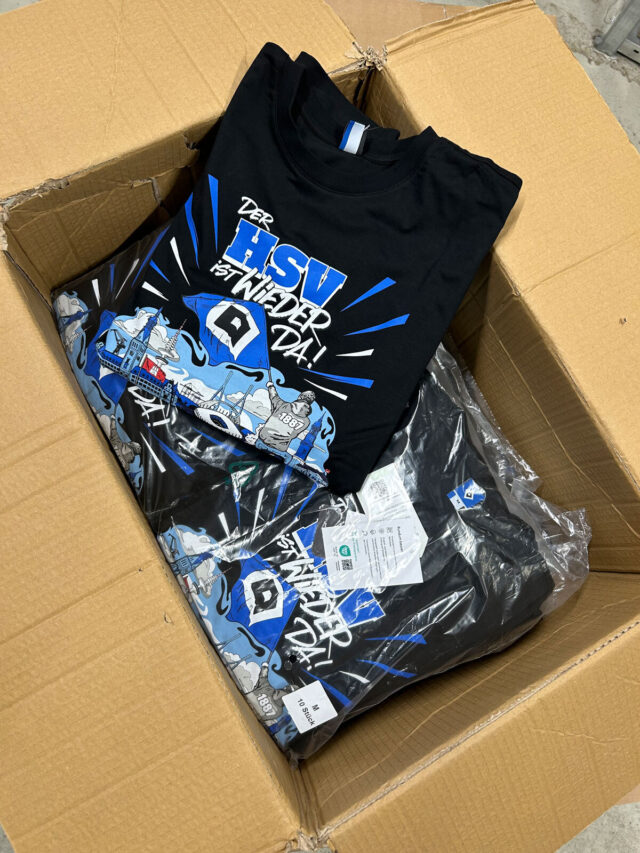 Auf dem Bild ist ein geöffneter Versandkarton zu sehen, in dem mehrere schwarze T-Shirts mit dem Aufdruck „Der HSV ist wieder da!“ liegen. Die Shirts zeigen ein farbenfrohes Motiv in Blau, Weiß und Grau mit HSV-Logo und Hamburger Wahrzeichen. Einige der T-Shirts sind gefaltet und in Polybags verpackt. Es handelt sich um das offizielle Aufstiegsshirt des Hamburger SV.