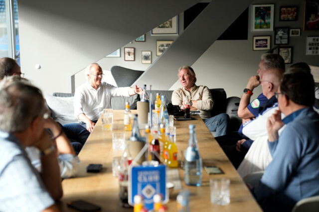 Das Bild zeigt einen Raum mit einem Tisch in der Mitte, auf dem viele Getränke stehen. Um den Tisch herum sitzen sieben Personen mit dem Blick auf zwei Männer an der Stirnseite. Die Männer sind Horst Hrubesch und Bernd Wehmeier. Sie sitzen auf einem grauen Sofa und unterhalten sich mit den Personen im Raum.