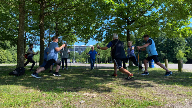 Das Bild zeigt eine Gruppe älterer Menschen in Sportkleidung unter Bäumen, die mit Nordic-Walking-Stöckern eine Gymnastikübung machen. Im Hintergrund ist das Volksparkstadion zu sehen.