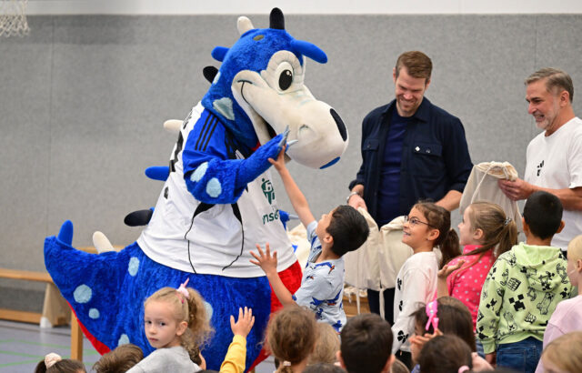Das Foto zeigt Dino Hermann in einer Sporthalle. Um ihn herum sind Kinder, die mit ihm abklatschen. Im Hintergrund sind die beiden Stiftungsvorstände zu sehen.