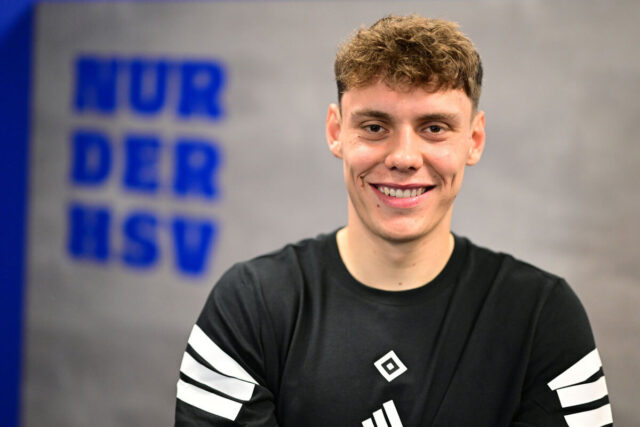Das Bild zeigt Nicolai Remberg, einen Spieler der HSV-Männer, vor einer grauen Wand, auf der links im Hintergrund in Blau „Nur der HSV“ steht. Remberg trägt ein schwarzes T-Shirt mit der HSV-Raute in der Mitte. Seine Haare sind braun, kurz und lockig. Er lächelt breit in die Kamera.