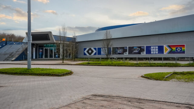 Auf dem Bild ist das HSV-Zentrum am Volkspark zu sehen. Links führt eine breite Treppe nach oben, daneben befindet sich ein verglaster Eingangsbereich. Entlang der langen Fassade hängen mehrere große Banner – darunter die HSV-Raute, der Slogan „Nur der HSV“, der Slogan „Love Hamburg hate racism“ sowie ein Banner in den Farben der Progressive Pride Flag mit Raute. Der weitläufige Vorplatz aus Pflastersteinen und kleinen begrünten Flächen wirkt ruhig und menschenleer.
