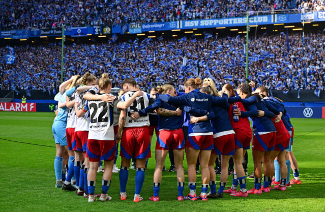 Das Foto zeigt die HSV-Frauenmannschaft, die sich vor dem Spiel in einem Kreis auf dem Spielfeld versammelt hat. Die Spielerinnen stehen eng beieinander, haben die Arme umeinandergelegt und stimmen sich gemeinsam ein. Im Hintergrund ist das Stadion mit zahlreichen HSV-Fans zu sehen, die in blauer Kleidung und mit Schals ihr Team unterstützen.