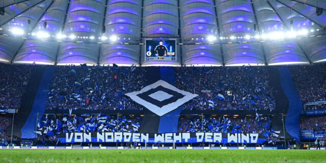Das Bild zeigt den Blick vom Rasen auf die Nordtribüne. Es ist während des HSV-Heimspiels gegen Ulm in der Saison 2024/25 entstanden und bildet die Fan-Choreo ab. Links, in der Mitte und rechts hängen von oben nach unten schwarz-blaue Banner herab. In der Mitte der Tribüne wird die Raute in Schwarz und Weiß hochgehalten. Drum herum halten die Fans blau-weiß-schwarze Fahnen hoch. Am unteren Rand steht auf einem großen Banner: „Von Norden weht der Wind.“