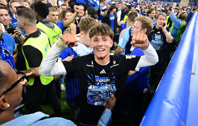 Mitten im Jubel der Fans steht ein strahlender HSV-Spieler, Otto Stange, der beide Fäuste triumphierend in die Luft hebt. Er trägt das schwarze Aufstiegsshirt mit dem Aufdruck „Der HSV ist wieder da!“ und wird von begeisterten Menschen umringt, die den Erfolg gemeinsam feiern. Die Stimmung im Stadion ist ausgelassen und voller Emotionen.
