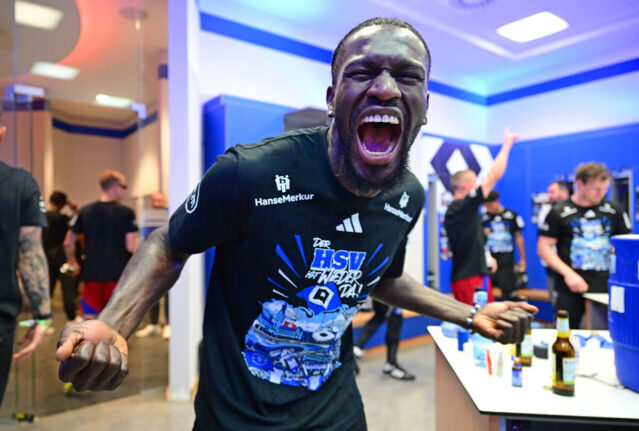Auf dem Bild ist HSV-Spieler Bakery Jatta in der Umkleidekabine zu sehen. Mit weit geöffnetem Mund und geballten Fäusten jubelt er voller Energie über den Aufstieg. Er trägt das schwarze Aufstiegsshirt mit dem Aufdruck „Der HSV ist wieder da!“. Im Hintergrund feiern weitere Teammitglieder ausgelassen mit.