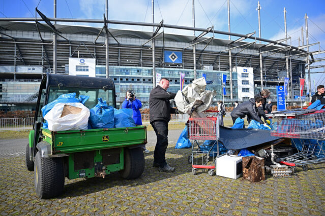 Das Foto zeigt ein Arbeitsfahrzeug auf dem Parkplatz vor dem Volksparkstadion, auf das Müllsäcke geladen wurden. Daneben stehen Personen, die volle Müllsäcke sortieren.