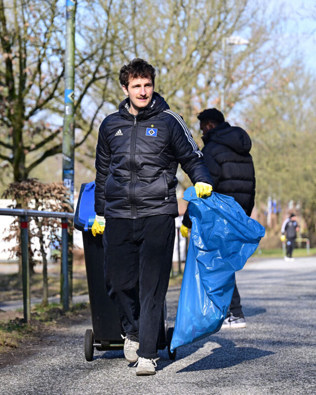 Das Bild zeigt einen jungen Mann draußen. Er trägt einen blauen Müllsack in der linken Hand und zieht mit der rechten Hand eine Mülltonne hinter sich her. Er trägt gelbe Gummihandschuhe und eine HSV-Winterjacke.