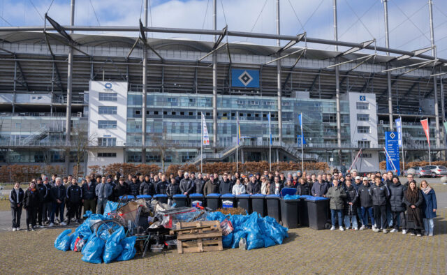 Das Foto zeigt eine große Gruppe von Menschen, die hinter einer Reihe von Mülltonnen und aufeinandergestapelten Müllsäcken und Sperrmüll stehen. Im Hintergrund ist das Volksparkstadion.