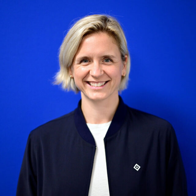 Auf dem Foto ist Laura Ludwig, Vizepräsidentin des Hamburger Sport-Verein e.V., zu sehen. Sie lächelt in die Kamera, hat kurze blonde Haare und steht vor einem blauen Hintergrund. Sie trägt eine dunkelblaue Jacke mit einem kleinen weißen Logo sowie ein weißes T-Shirt.