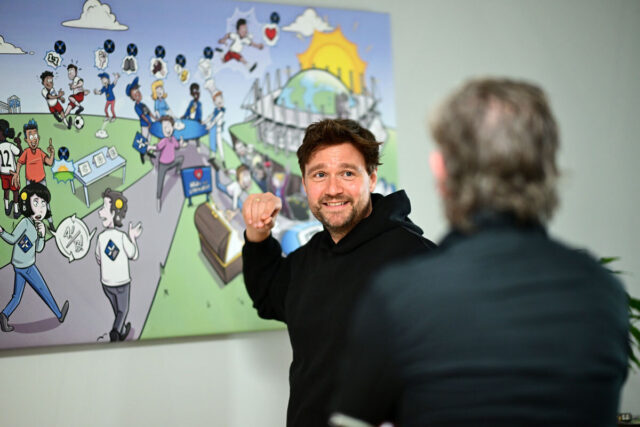 Das Foto zeigt Frank Weiland vor einem Wandbild. Das Bild ist eine Art Comicdarstellung des Volksparkstadions. Im Vordergrund ist verschwommen der Rücken einer Person zu sehen, mit der Frank Weiland sich unterhält.