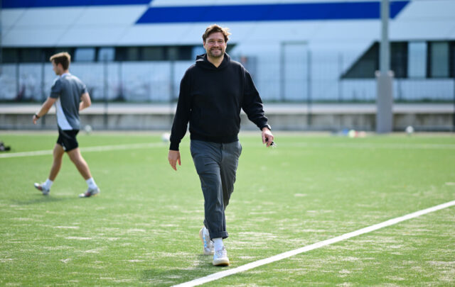 Das Foto zeigt einen Mann, der auf einem der HSV-Trainingsplätze auf die Kamera zuläuft. Der Mann ist der Sportpsychologe Frank Weiland. Im Hintergrund zu sehen sind die Alexander-Otto-Akademie sowie eine Person mit Fußballschuhen und grauem Trainer-Shirt.