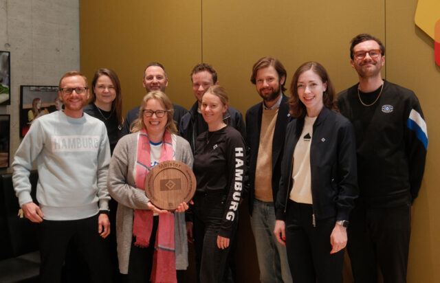 Auf dem Bild steht eine Gruppe von neun Personen zusammen und lächelt in die Kamera. In der Mitte hält eine Frau eine hölzerne Meisterplakette mit dem HSV-Logo in den Händen. Die Gruppe trägt überwiegend Freizeitkleidung in Blau-, Grau- und Schwarztönen, teils mit HSV- oder Hamburg-Aufdruck. Der Hintergrund ist schlicht mit einer goldfarbenen Wand und modernen Lichtelementen gestaltet. Die Stimmung wirkt fröhlich und stolz.