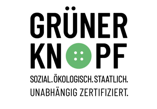 Auf dem Bild ist das Logo des staatlichen Textilsiegels „Grüner Knopf“ zu sehen. In der Mitte steht groß der Schriftzug „GRÜNER KNOPF“, wobei das „O“ durch einen grünen Knopf mit vier Löchern ersetzt ist. Darunter steht der Slogan: „Sozial. Ökologisch. Staatlich. Unabhängig zertifiziert.“
