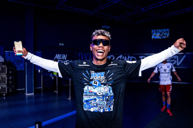 Auf dem Bild ist Ransford Königsdörffer zu sehen, der beide Arme ausbreitet und breit lächelt. Er trägt eine Sonnenbrille und das schwarze Aufstiegsshirt mit dem Aufdruck „Der HSV ist wieder da!“, das in Blau, Weiß und Grau gestaltet ist. In der einen Hand hält er ein Smartphone. Im Hintergrund sind weitere Spieler und blau beleuchtete Wände mit HSV-Schriftzügen zu erkennen.