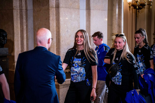 In einem repräsentativen Gebäude mit Marmorsäulen begrüßen HSV-Spielerinnen den Bürgermeister. Eine der Spielerinnen, die das schwarze Aufstiegsshirt mit dem Aufdruck „Der HSV ist wieder da!“ trägt, lächelt freundlich beim Händeschütteln. Im Hintergrund sind weitere Spielerinnen zu sehen, die ebenfalls das Shirt tragen und blaue Jacken in den Händen halten.